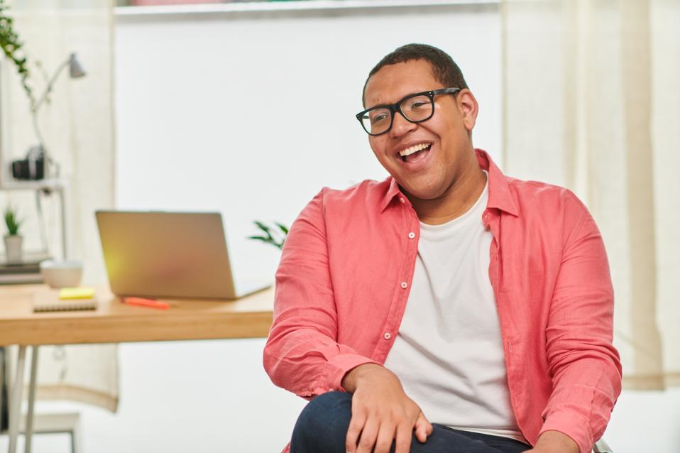 Smiling man in glasses, an adult with disabilities, sits laughing in a bright office space