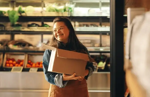 Smiling young woman with Down syndrome carrying a box in a grocery store, an adult with disabilities at work